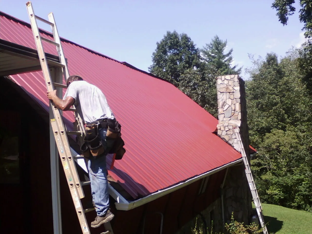 Roofer on a ladder beside a red metal roof and stone chimney — Soffit & Fascia Repair by Roofing Brothers in Dothan